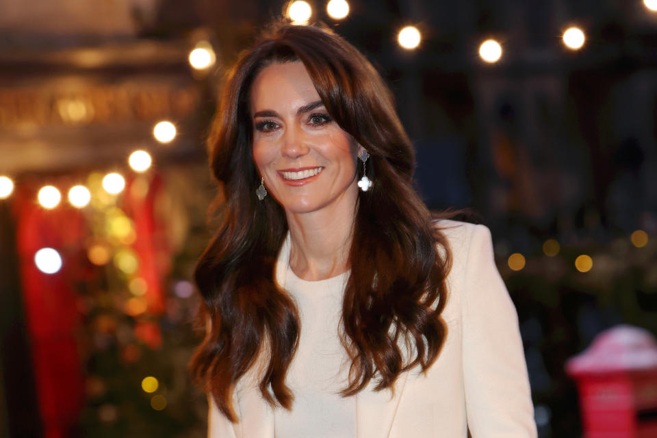 Kate Middleton smiles while wearing a blazer and earrings at an outdoor evening event with string lights in the background