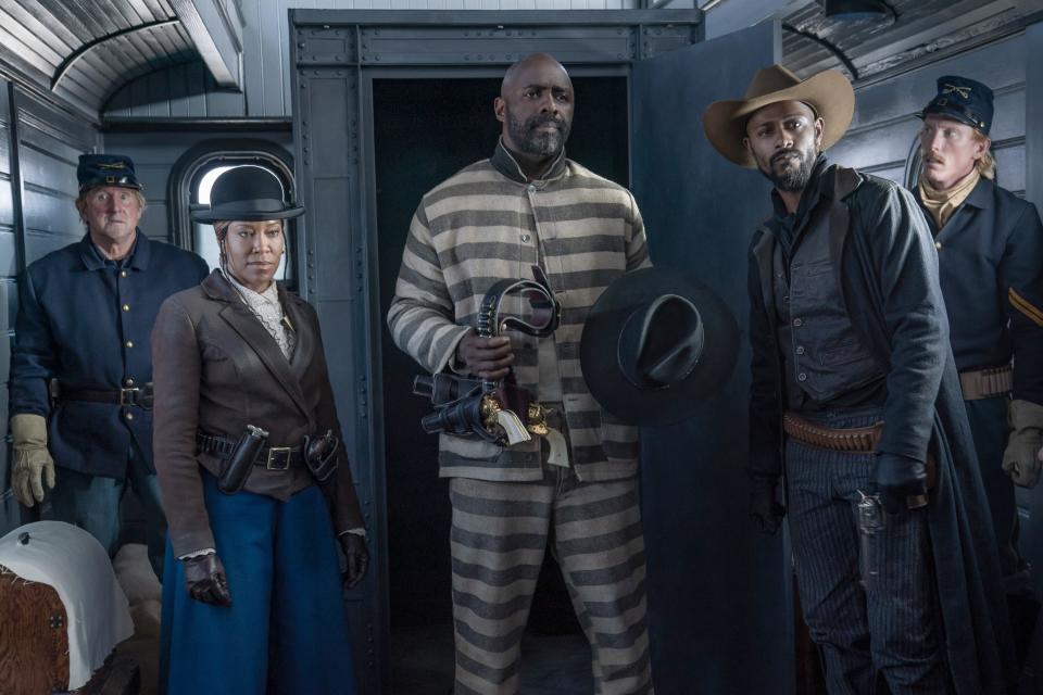 A Black man stands in a prison uniform, he holds a black hat and gun. A Black woman and Black man both in hats and guns stand on either side.