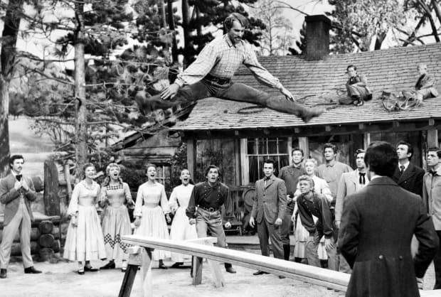 Caleb Pontipee, played by American jazz and ballet dancer Matt Mattox (1921 - 2013) , leaps above a sawhorse in 'Seven Brides For Seven Brothers', directed by Stanley Donen, 1954. Amongst those looking on are Tommy Rall (6th from left) as Frank Pontipee, and Russ Tamblyn (9th from left) as Gideon Pontipee.<p>Silver Screen Collection/Getty Images</p>