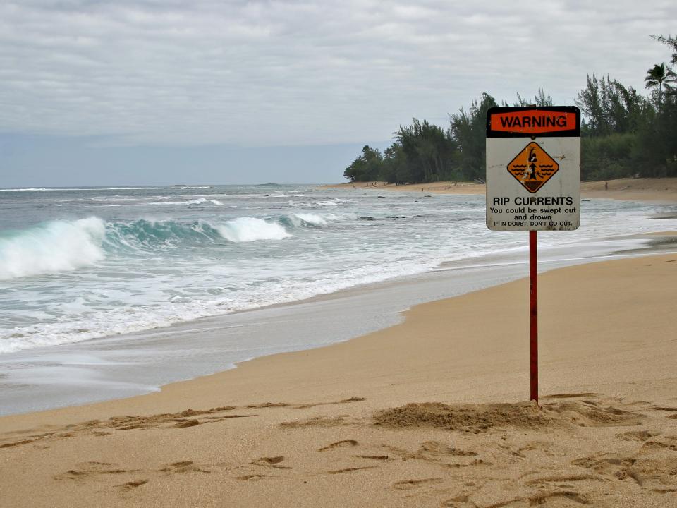 A rip current warning sign on a beach near Kauai, Hawaii, and the sign says: "Rip Currents - You could be swept out and drown - If in doubt, don't go out".