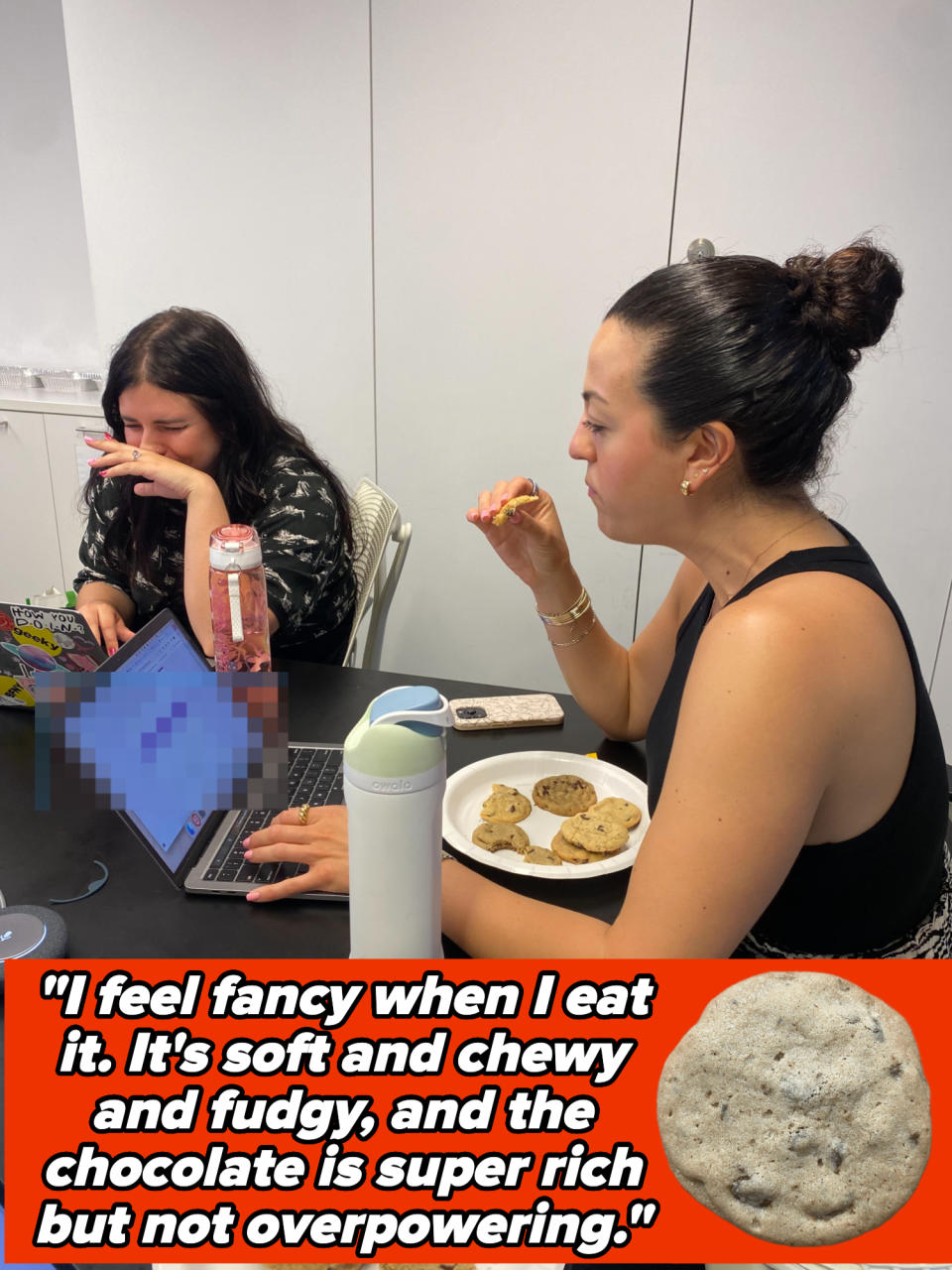 Two people sitting at a table with laptops and plates of cookies, eating and working. Cookies, a phone, and a water bottle are also on the table