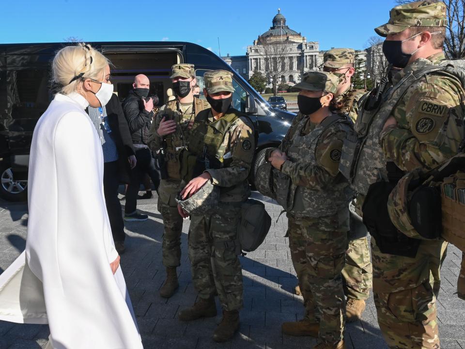 Gaga in a white cape and face mask meeting with soldiers in uniform outside.