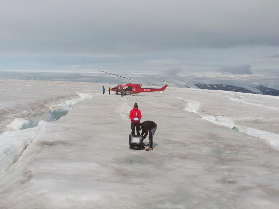 two scientists place samples in a box on a glacier while colleagues board a helicopter in the background
