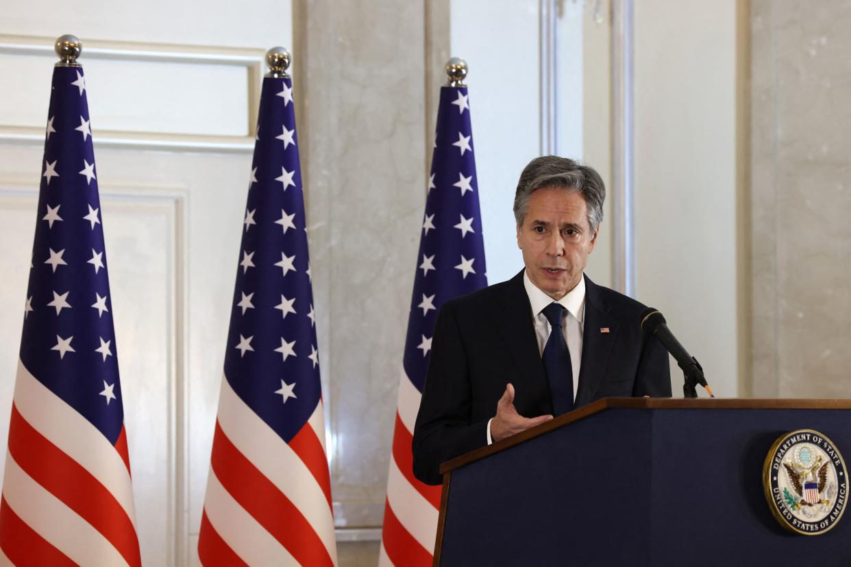 Secretary of State Antony Blinken at the microphone with three American flags behind him.