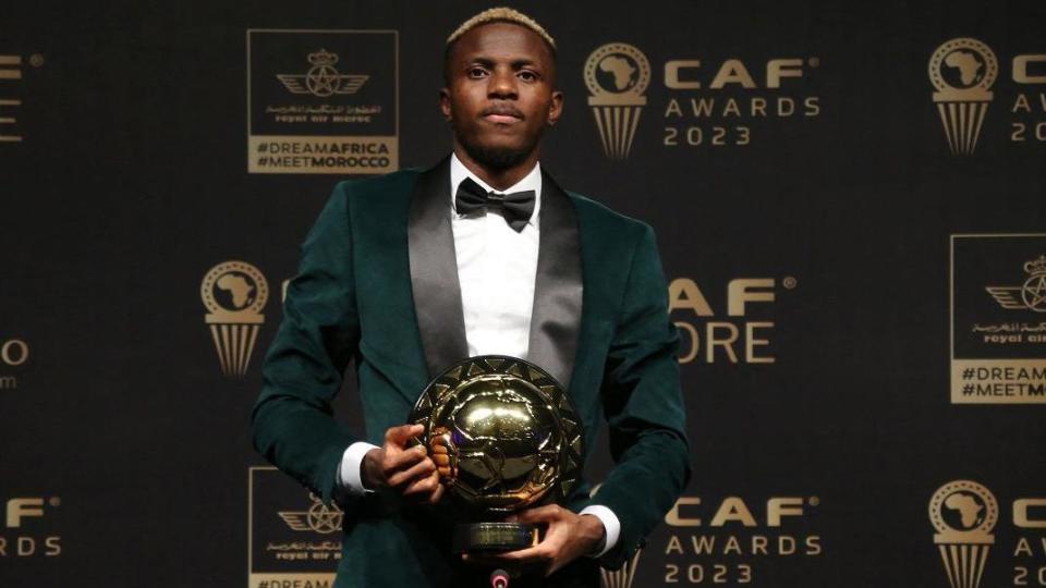 Victor Osimhen in green dinner jacket and black bow tie poses with his African Player of the Year trophy in front of boards promoting the Caf Awards 2023