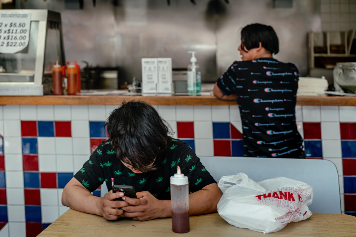 Un niño migrante, que trabaja el turno nocturno en una fábrica, mira su teléfono en un restaurante pequeño de comida rápida en el barrio de Little Village en Chicago, el 14 de abril de 2023. (Jamie Kelter Davis/The New York Times)