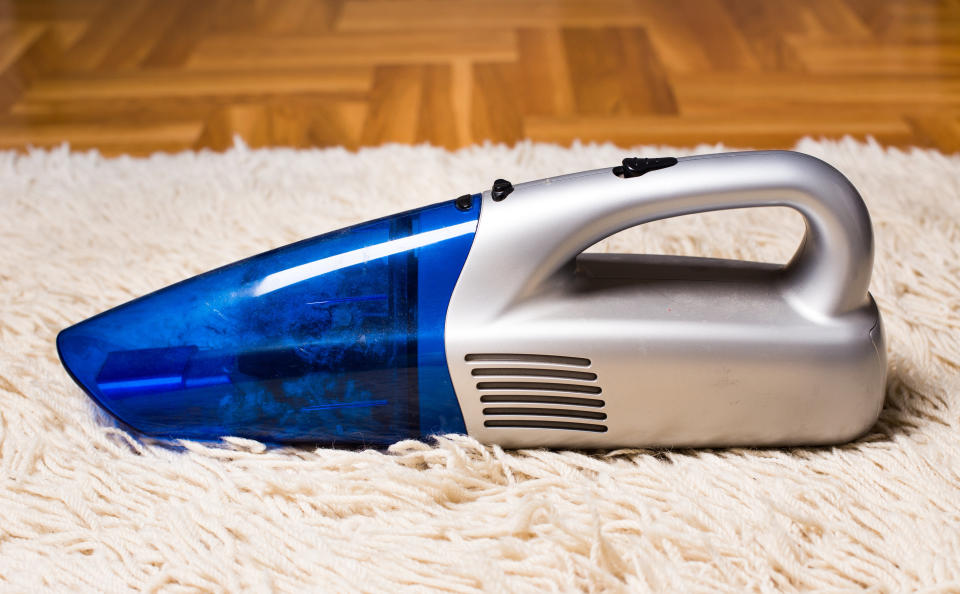 Handheld vacuum cleaner standing on white fluffy carpet with parquet in background. Housekeeping concept