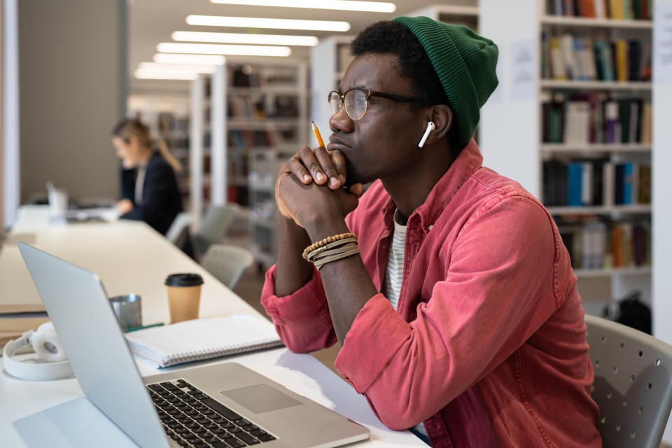 a man in a library with a laptop, headphones, and coffee, thoughtfully looking out of the window