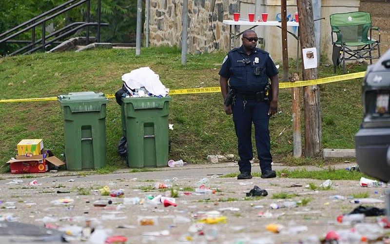 A police officer stands in the area of a mass shooting. Behind him are trash cans and police caution tape, and on the ground in front of him are scattered plastic bottles and cups.