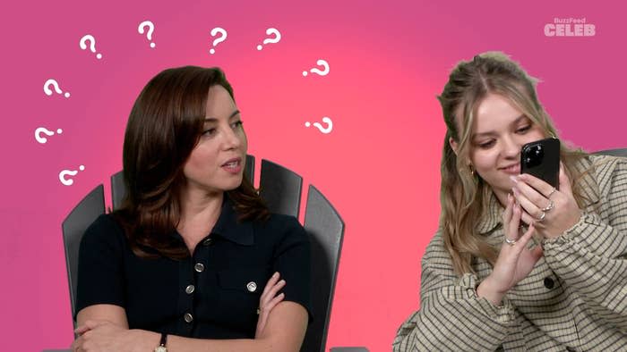 Aubrey Plaza and Maisy Stella sit in chairs. Aubrey looks at Maisy, who is holding a phone and smiling. Question marks are above Aubrey’s head