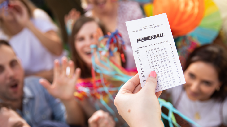 Photo of Powerball ticket in hand in the foreground, with people celebrating with streamers blurred in the background.