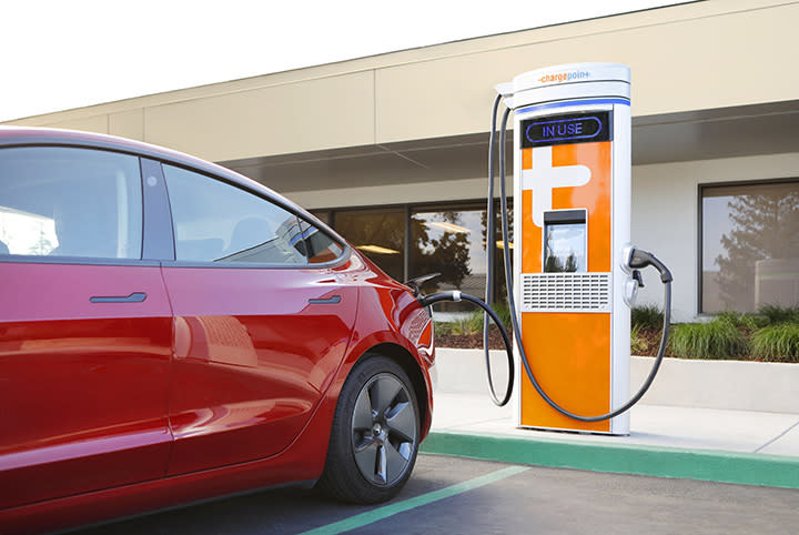 ChargePoint marketing image of an electric charging station. A red car is plugged into a (primarily) orange charging station sitting atop a curb in front of a building.