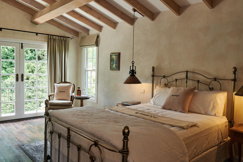 A room with cream rendered walls, exposed beams, French doors, brass bed, armchair and brass lighting