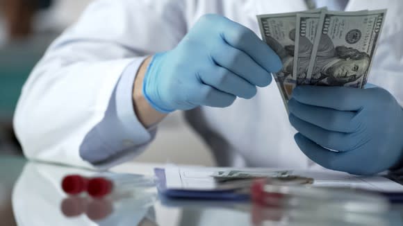 Person wearing a lab coat and gloves counting hundred dollar bills.