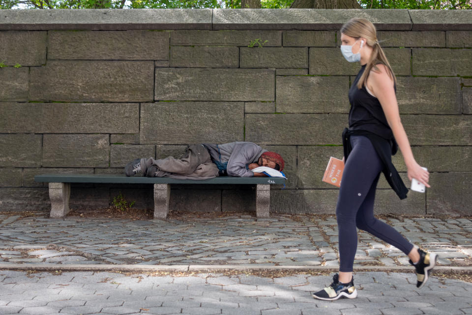 NEW YORK, NEW YORK - MAY 03: A woman wearing a mask walks by a homeless person sleeping on a bench as temperatures rise amid the coronavirus pandemic on May 3, 2020 in New York City. COVID-19 has spread to most countries around the world, claiming over 248,000 lives and infecting over 3.5 million people. (Photo by Alexi Rosenfeld/Getty Images)