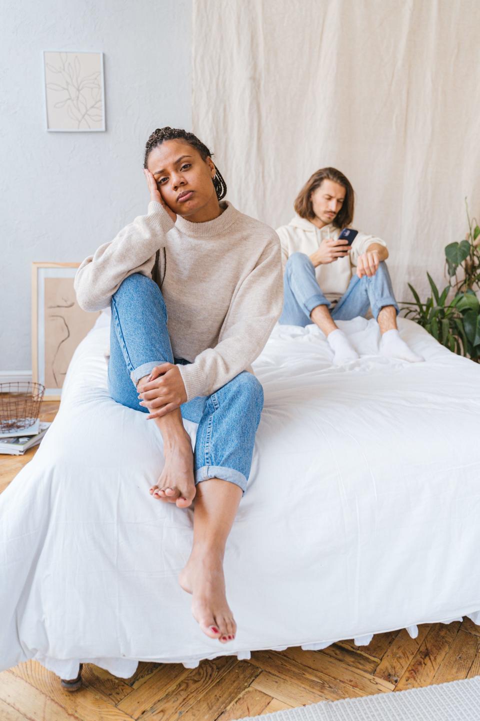Frustrated Black woman sit on the edge of the bed while her partner stares at his phone