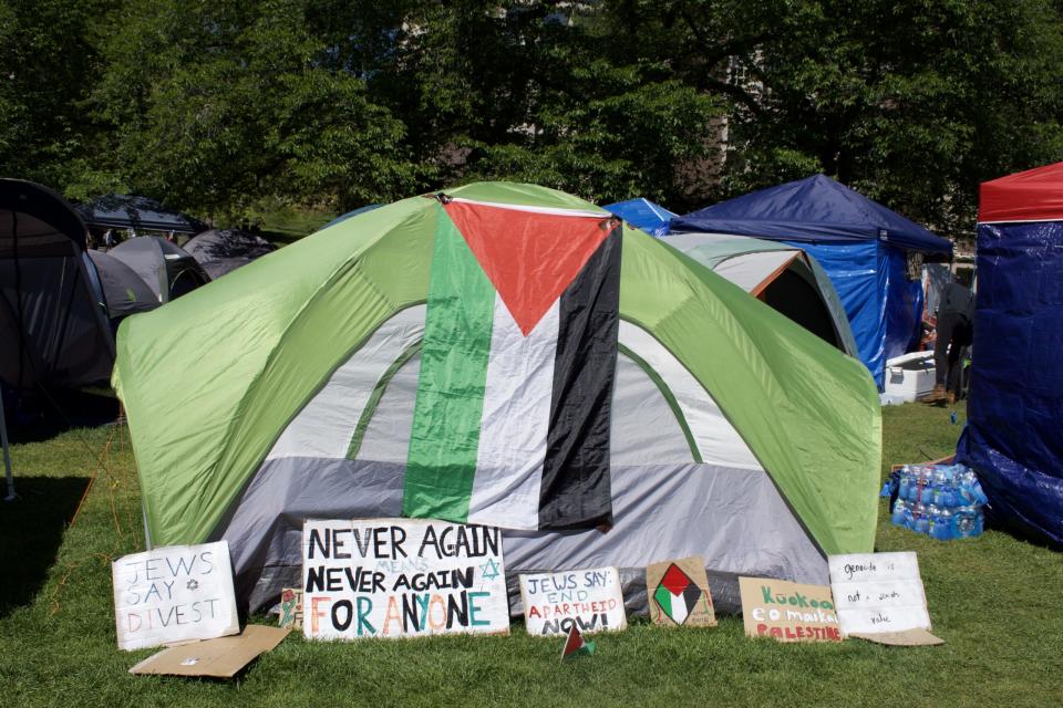 A Palestinian flag hangs in front of a tent. Below, signs say things like "never again means never again for anyone" and "Jews say divest." 
