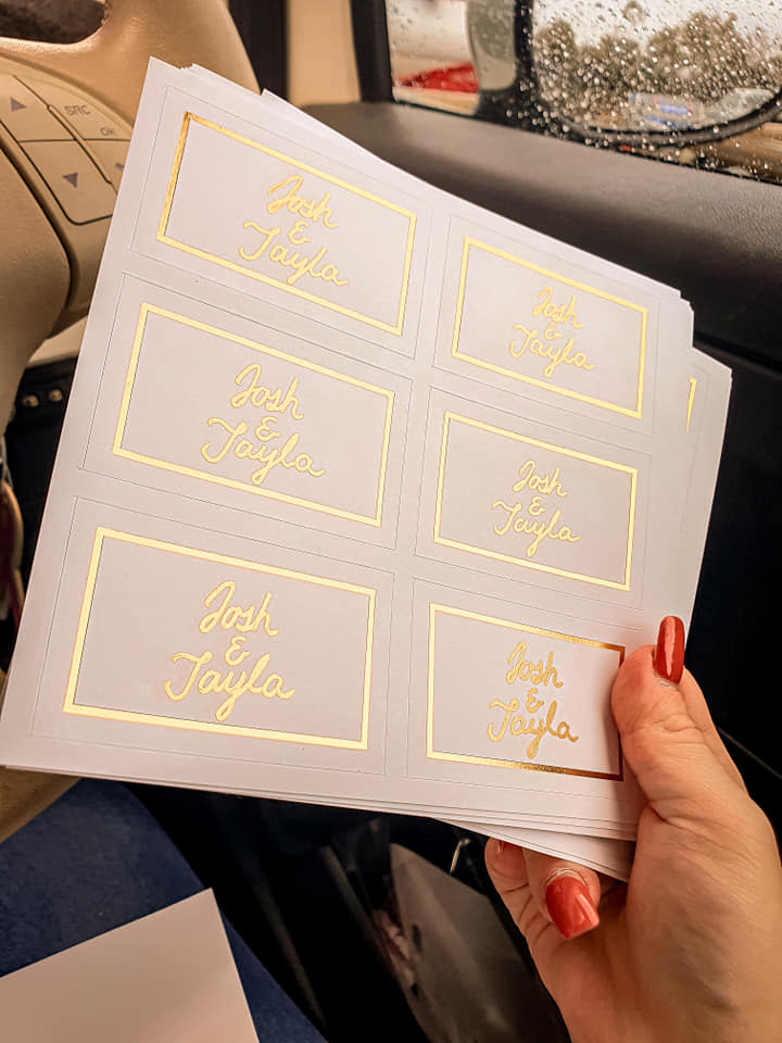 Woman's hand holding sheets of white labels that read 'Josh & Tayla' in gold pen