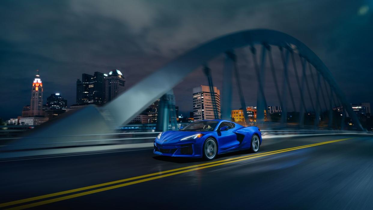 front 34 view of 2024 chevrolet corvette e ray 3lz coupe in riptide blue, driving over a bridge in front of a city pre production model shown actual production model may vary model year 2024 corvette e ray available 2023