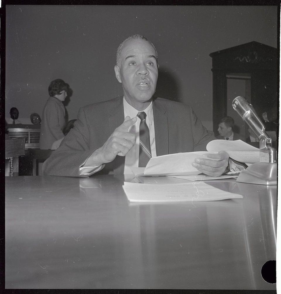 roy wilkins sits at a table and looks ahead while speaking and holding papers in one hand, he wears a suit jacket, tie and collared shirt, two people and a doorway are in the background