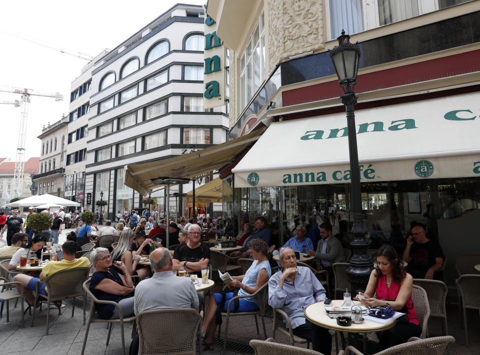 In this photo taken on Wednesday, June 5, 2019, people sit at a cafe on Vaci Street in downtown Budapest. A tourism boom in the Hungarian capital has led to major congestion on the river flowing through the city, with sightseeing boats and floating hotels competing for better positions in front of spectacular neo-Gothic buildings, ornate bridges and churches lining the Danube. (AP Photo/Laszlo Balogh)