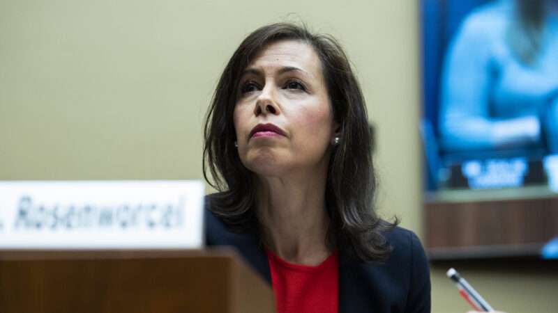 FCC Chairwoman Jessica Rosenworcel sits in a congressional hearing room behind a placard bearing her name, wearing a navy blazer and a red shirt and holding a mechanical pencil.