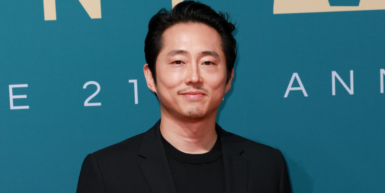 steven yeun smiles for the camera in a black tshirt and suit