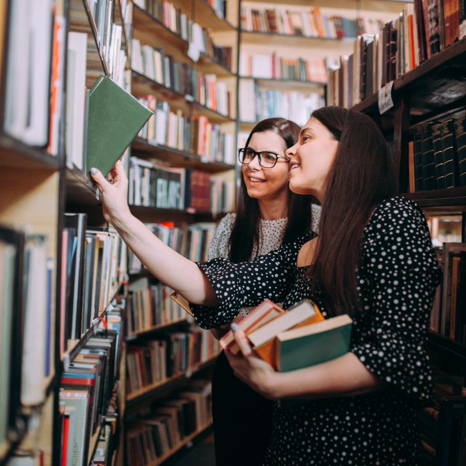 Two women stand in a library between shelves of books, smiling and selecting books to read. They both have long, dark hair and wear patterned dresses