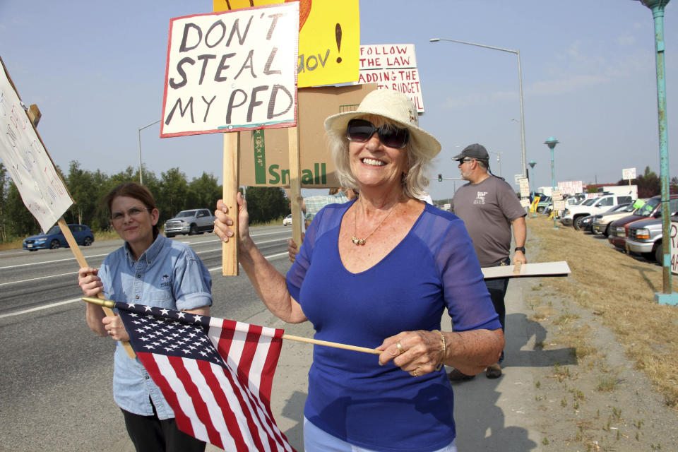 Katherine Hayes waves a flag and a sign urging Alaska lawmakers to fund a full oil wealth fund check, known locally as the PFD or Permanent Fund Dividend, Monday, July 8, 2019, in Wasilla, Alaska. Some Alaska lawmakers are meeting in Wasilla July 8 instead of Juneau, where state House and Senate leadership have decided to hold the special session, called to determine the amount of this year's check. (AP Photo/Mark Thiessen)