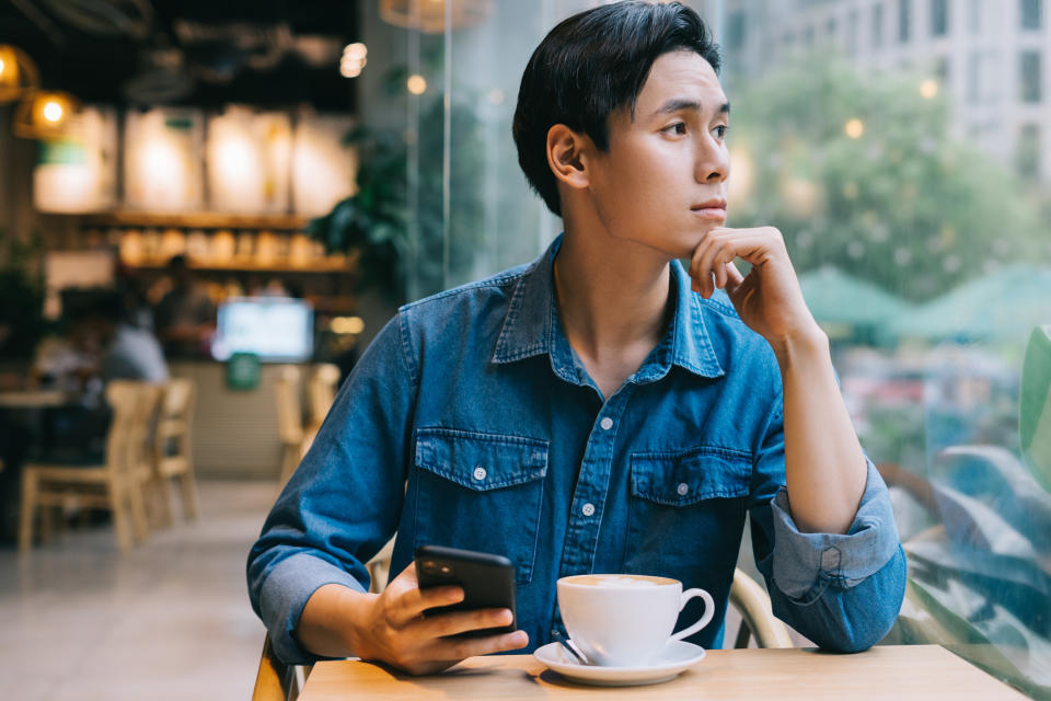 A person sits in a cafe, holding a smartphone in one hand and looking out the window thoughtfully. A coffee cup is on the table