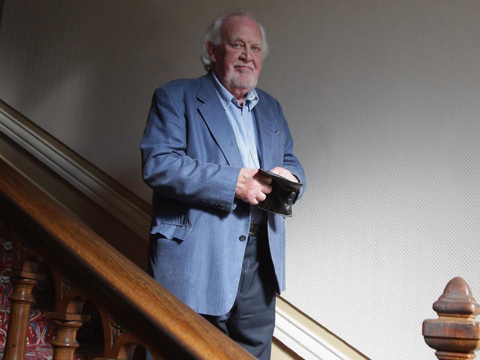 Joss Ackland standing on stairs wearing a blue coat