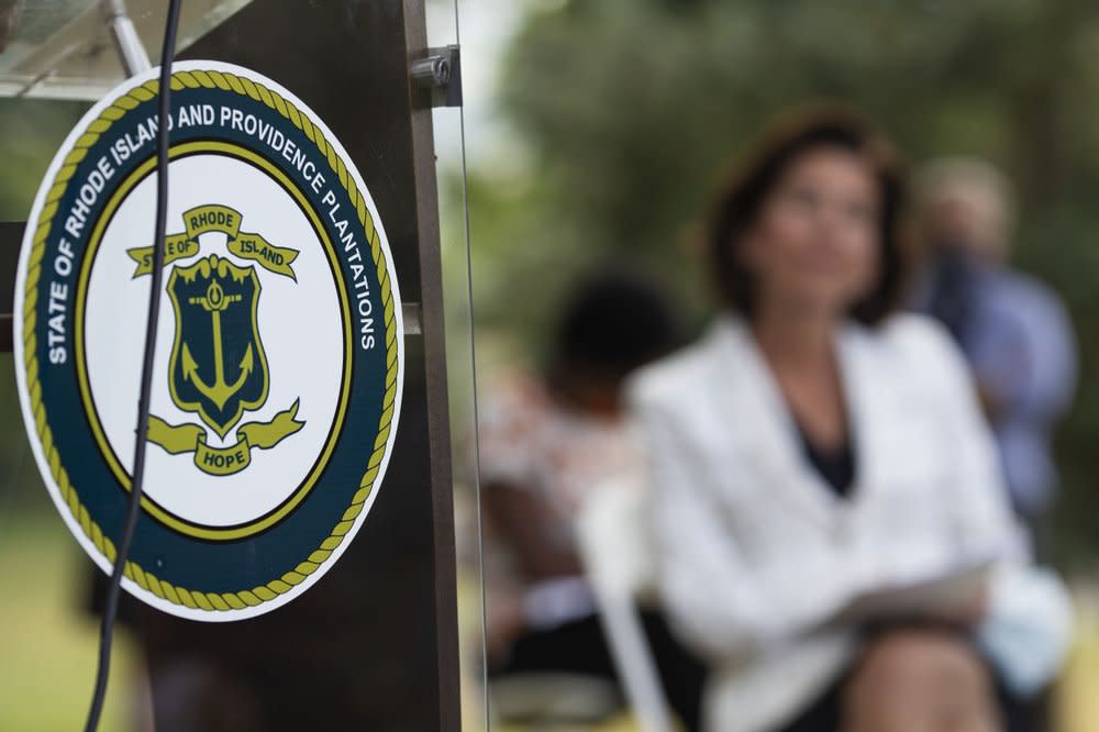 In this Monday, June 22, 2020, file photo, the seal of the State of Rhode Island decorates a podium as Gov. Gina Raimondo looks on at right during a news conference where she announced that she has signed an executive order to remove the phrase “Providence Plantations” in the state’s formal name from some official documents and executive agency websites in Providence, R.I. (AP Photo/David Goldman, File)