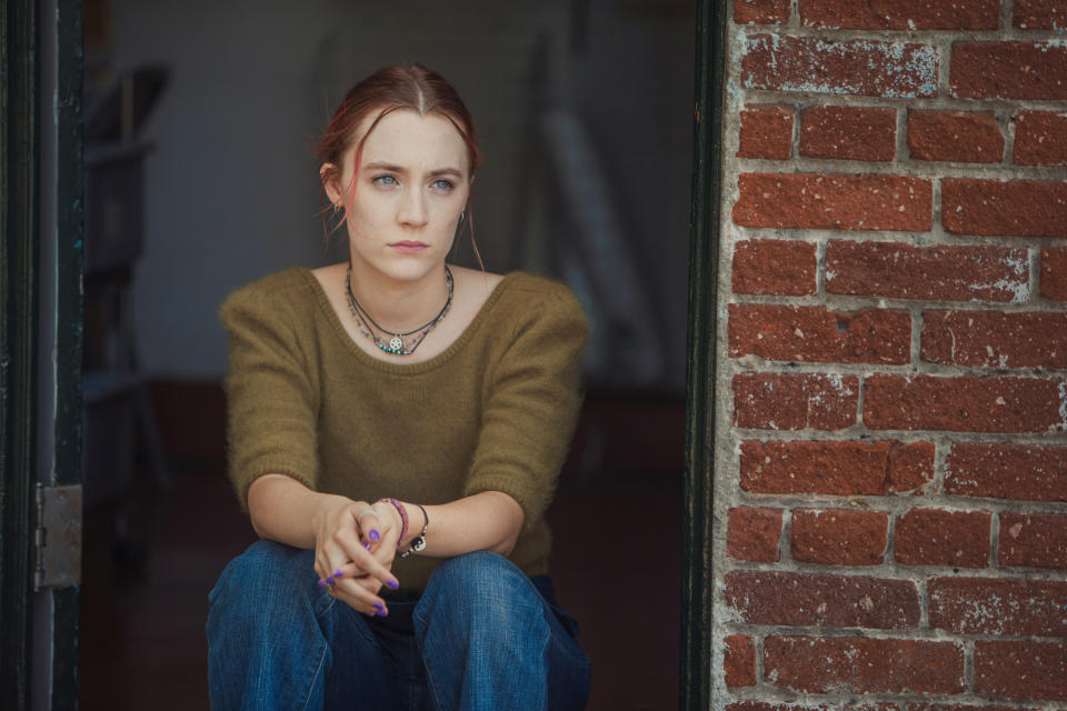 Lady Bird sitting by the window, wearing sweater and necklace, lost in thought