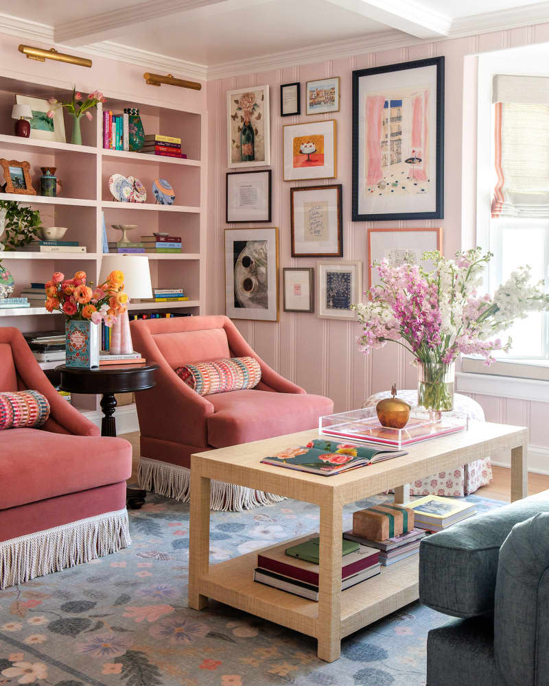 Two fringed pink chairs in front of light coffee table in pink library with gallery wall and built-in shelves.