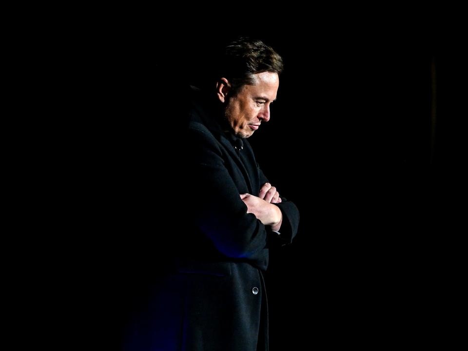 Elon Musk crossing his arms in front of a black background.