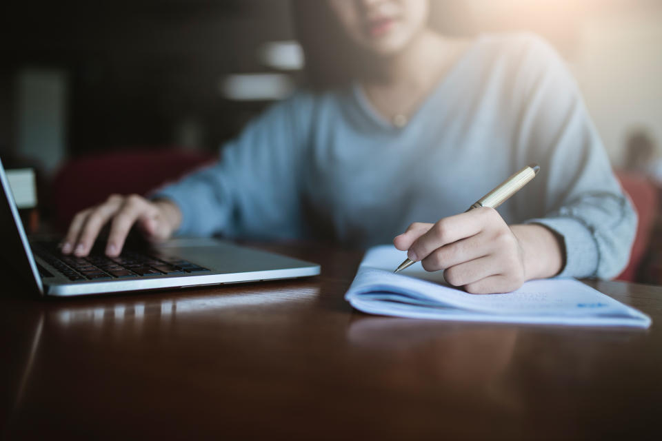 Midsection Of Student Writing On Book While Using Laptop In Library