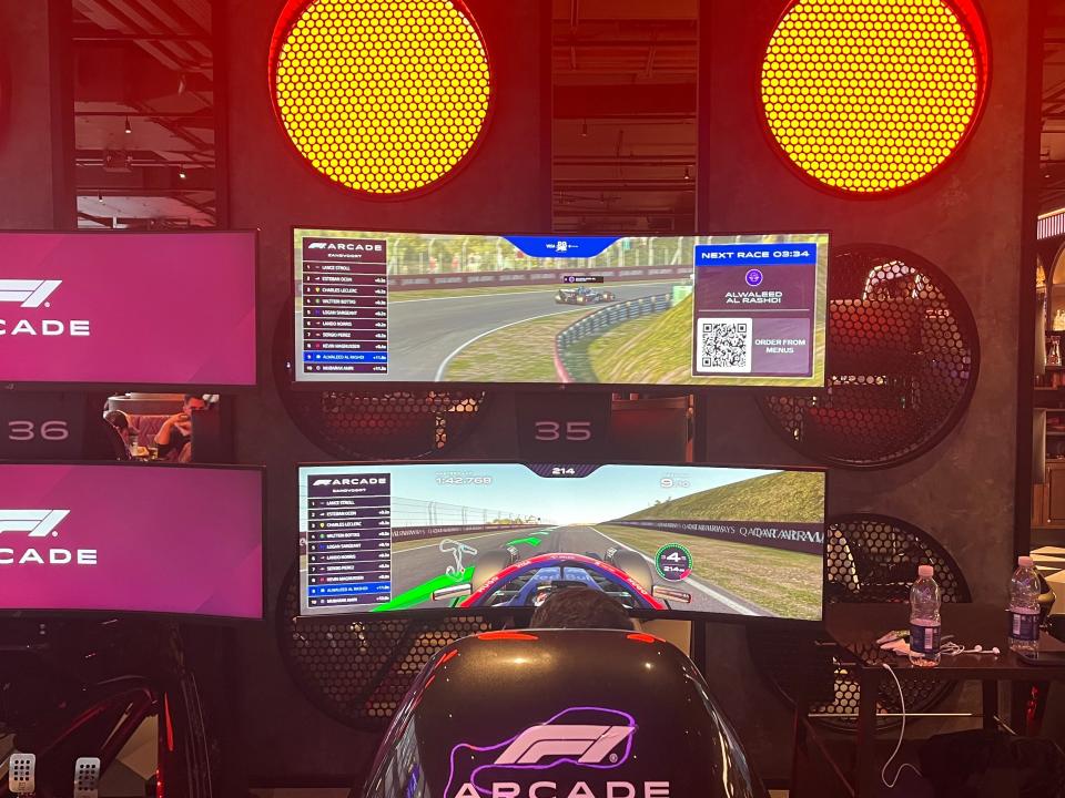 A driver behind the wheel of a driving simulator, with two screens showing a car on a track overhead.