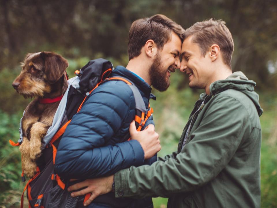 Two men press their noses together. One man carries a dog in a backpack.
