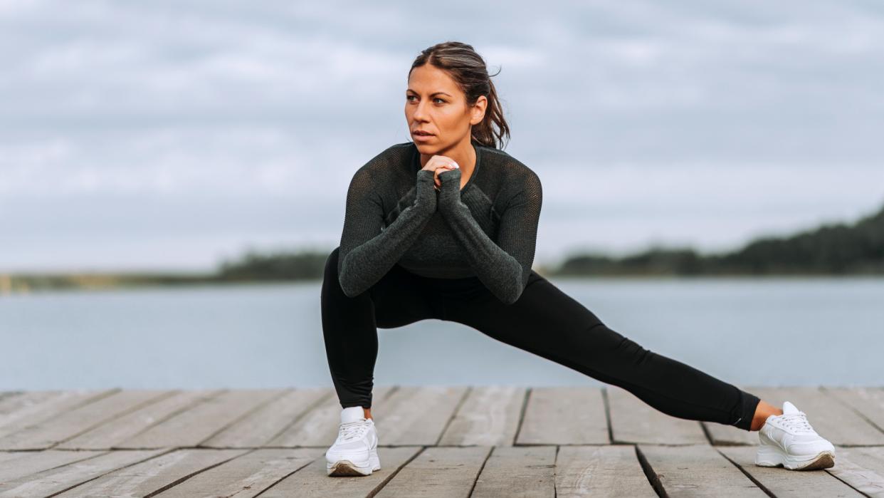  Woman performing a Cossack squat with right knee bent and left knee extended with backdrop of a river . 