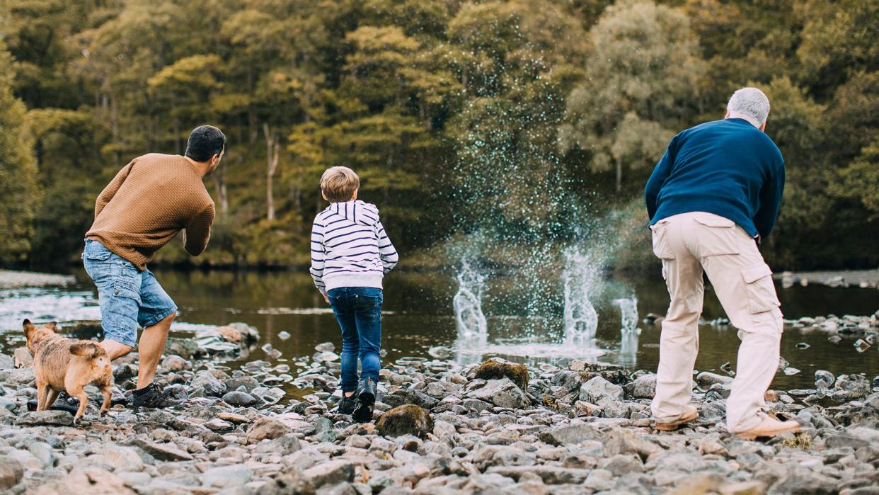 Little boy is skimming pebbles on a lake with his father and grandfather.