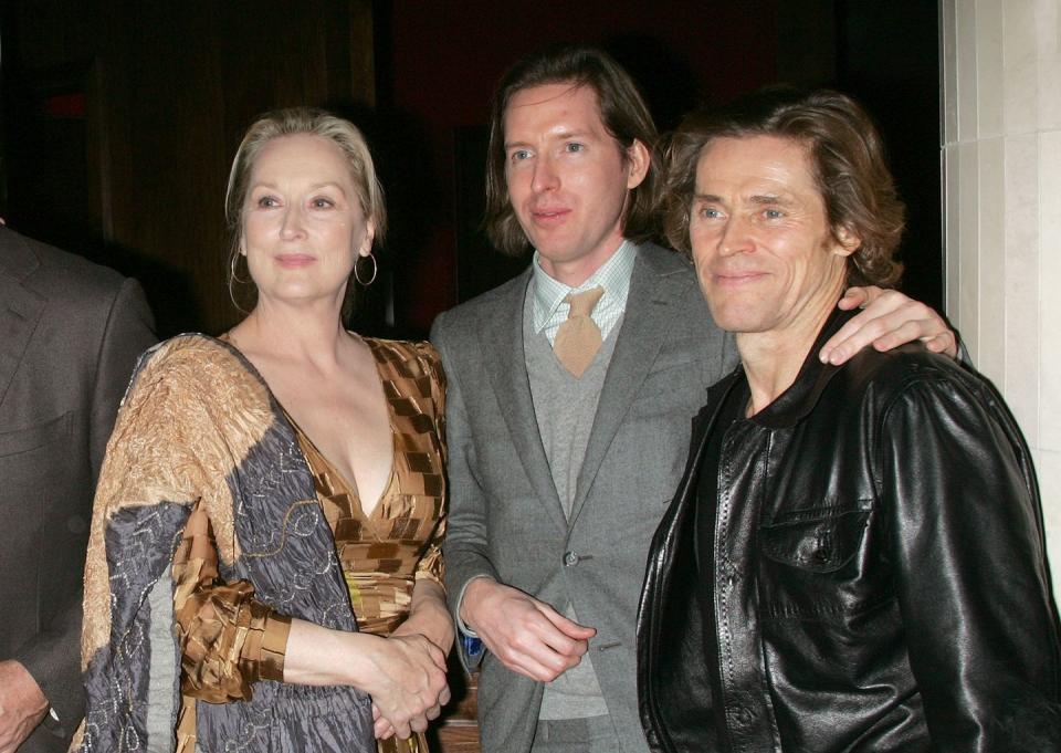 wes anderson, wearing a gray suit, puts his hand on the shoulder of willem dafoe, who wears a black leather jacket