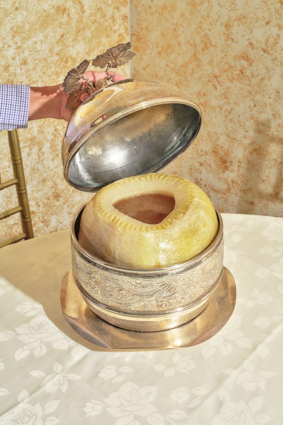 A hand lifts the lid of a round metal serving tureen containing a winter melon filled with soup