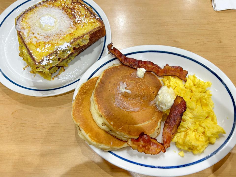 Plate of French toast next to a plate full of pancakes, bacon, and scrambled eggs on a wooden table at IHOP