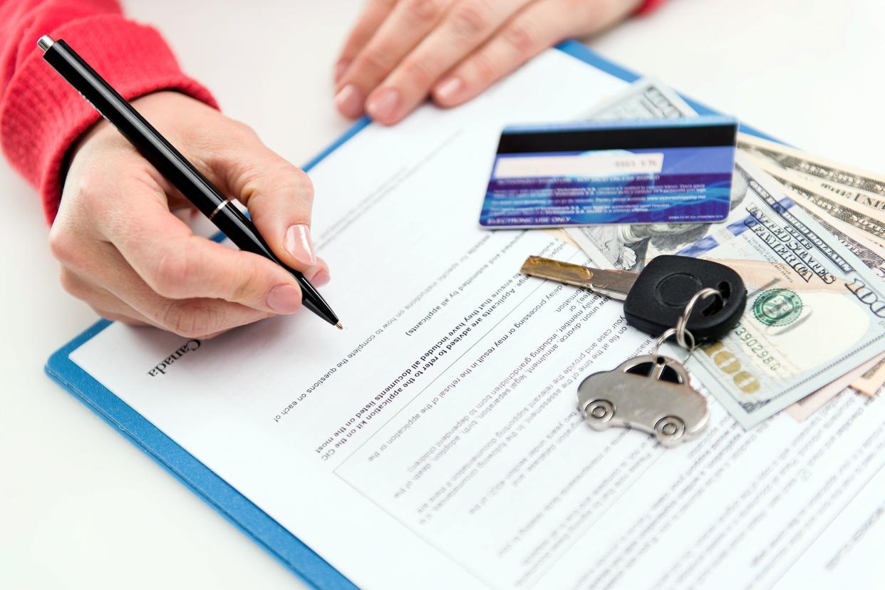 Closeup of woman signing a lease contact for car with credit card, us money, and car keys on contract on blue clipboard on white table