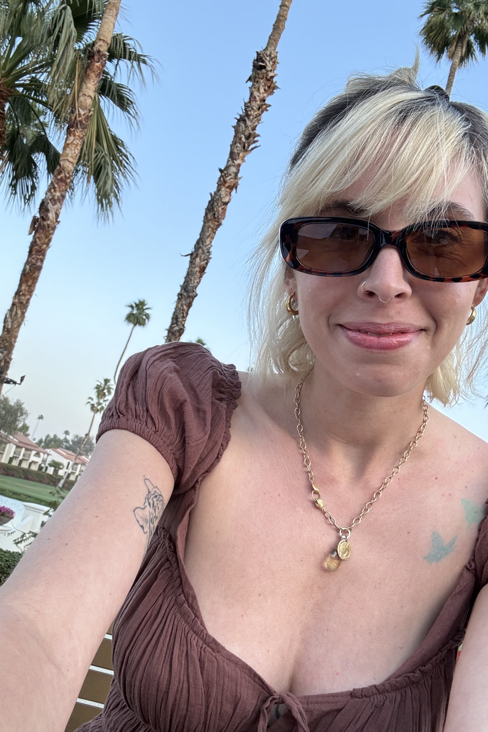 Woman in sunglasses and puffy-sleeved top takes a selfie with palm trees and a clear sky in the background