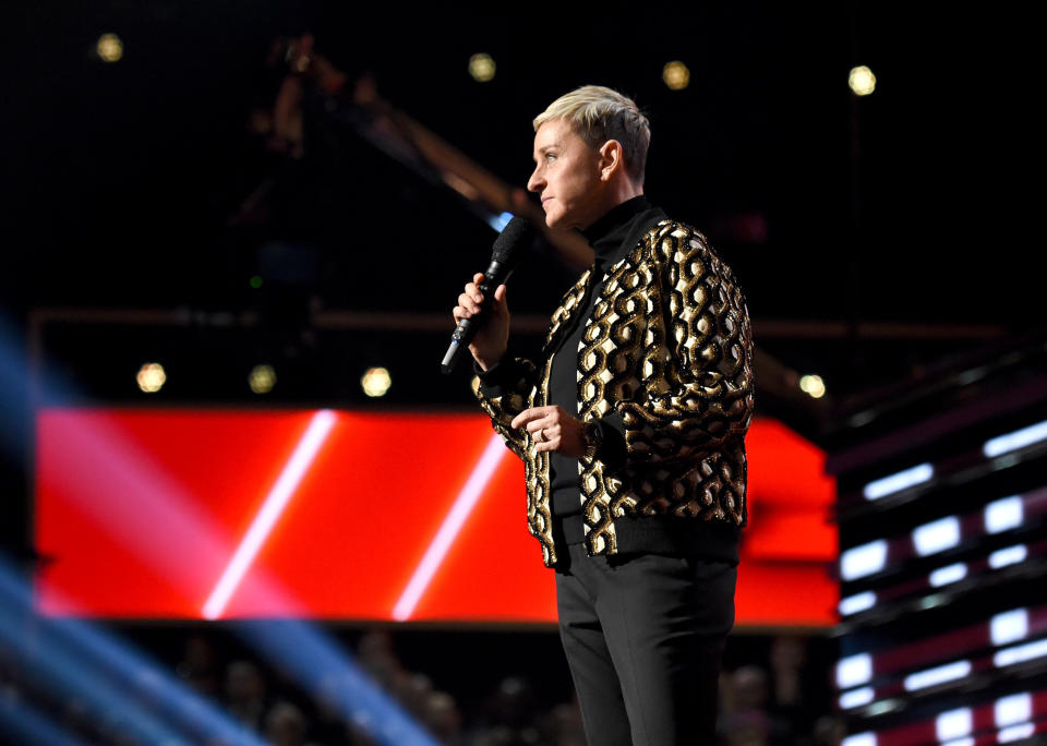 Ellen DeGeneres speaking on stage, wearing a black and gold patterned jacket