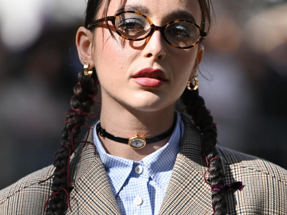A close up of a girl wearing blue eyeshadow, round glasses and her dark brown hair in two braids. She is wearing a watch as a choker necklace with a blue gingham shirt and checked blazer