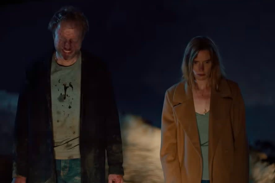 Two people, a man and a woman, stand in a dimly lit area. The man has a distressed look with a dirty face and clothes. The woman wears a coat and looks serious