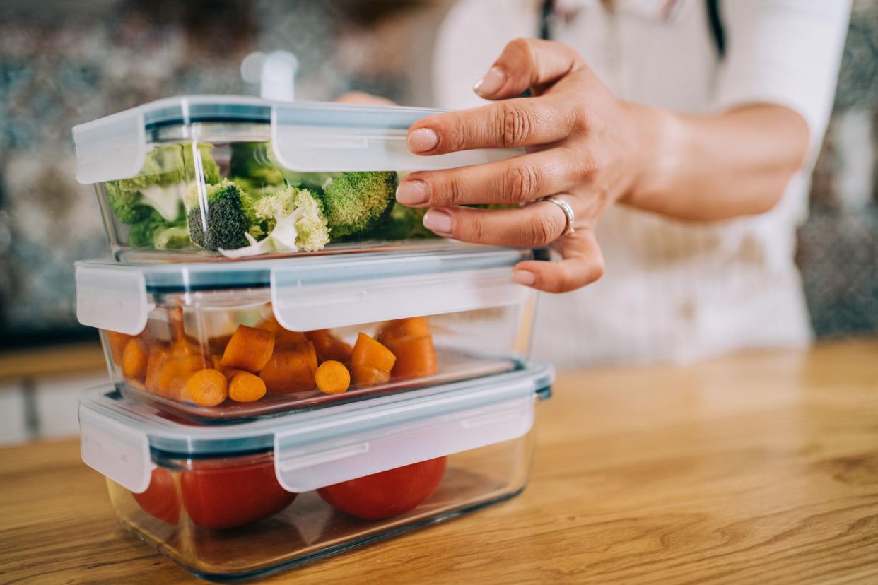 Close-up of female hands holding three glass tupperware containers with fresh raw vegetables to the left side, background is blurred