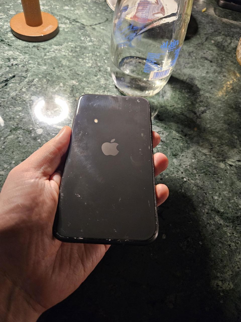 Hand holding a black iPhone with a visible Apple logo on a marble countertop next to a glass container